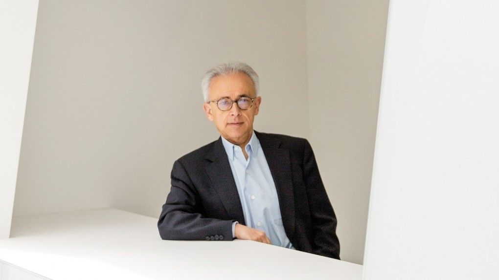Antonio Damasio leans on a white desk and looks into the camera.