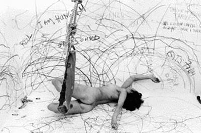Photograph of Carolee Schneemann naked drawing on a room-sized paper and attached to a swing.