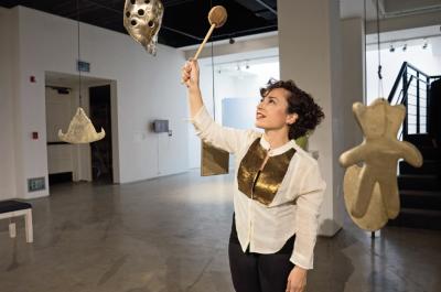 A photo of a woman with short dark curly hair wearing a white shirt and holding a mallet above her head to strike a hanging object.