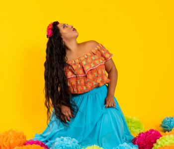Artist AAlicia Mullikin. She is against a yellow backdrop and surrounded by yellow, green, teal, and bright pink poms on the floor. She has long wavy hair spilling down the side of her shoulder and body and holding a teal dress, looking up to the right. Her top is a print containing the colors of the floor poms and is off the shoulders with ruffles across the top.