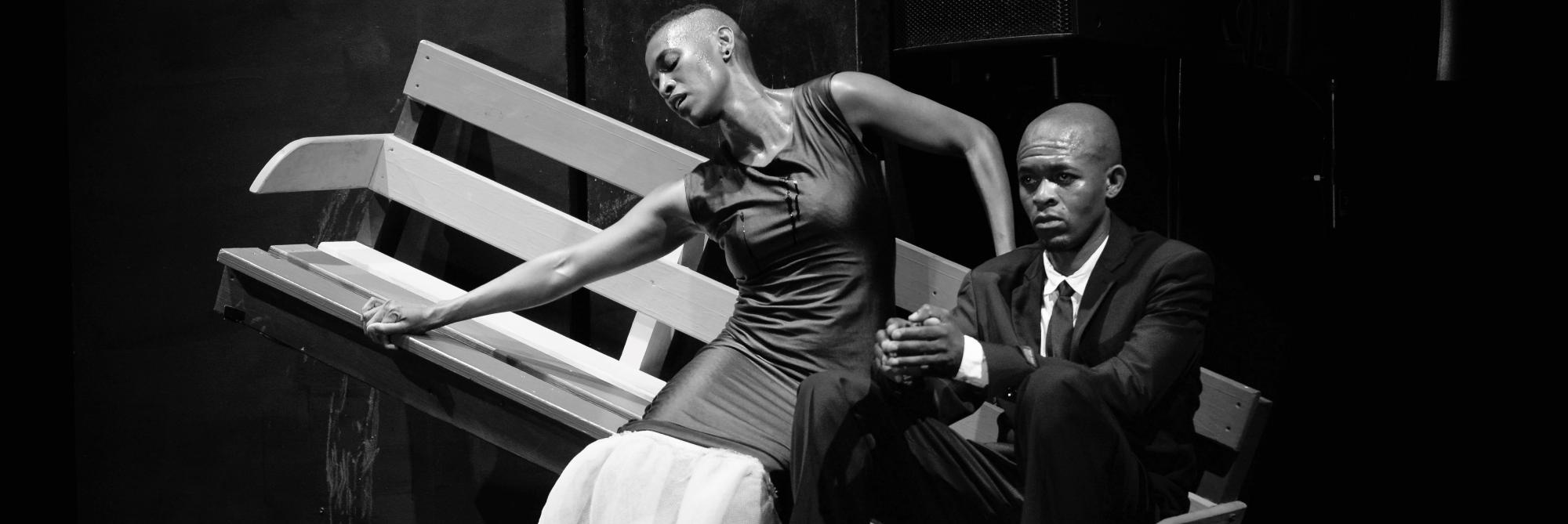 Photographed still from theatre production of Nyamza's De-Apart-Hate. Two people on a bench, one male-persenting in a suit looking off to the distance, one female-presenting in a dress reaching to the left. The bench is tilted so that the female-presenting performer appears to be sliding down to the right against the male-presenting performer.