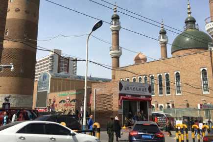A city street with multiple buildings, including a security checkpoint and a mosque nearby in the background