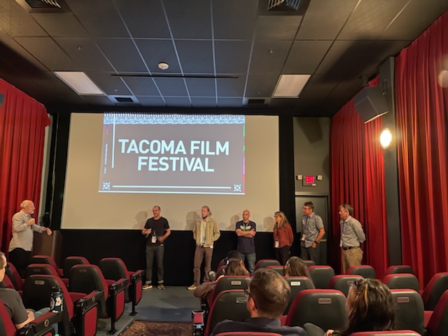 Six people stand on a stage while being interviewed by a seventh person. They all stand in front of a screen that reads Tacoma Film Festival and people sit in an audience in the foreground.