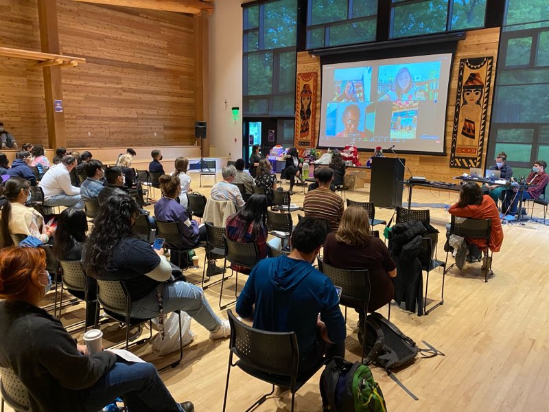 crowd seated and watching the panelists in person and on a projector