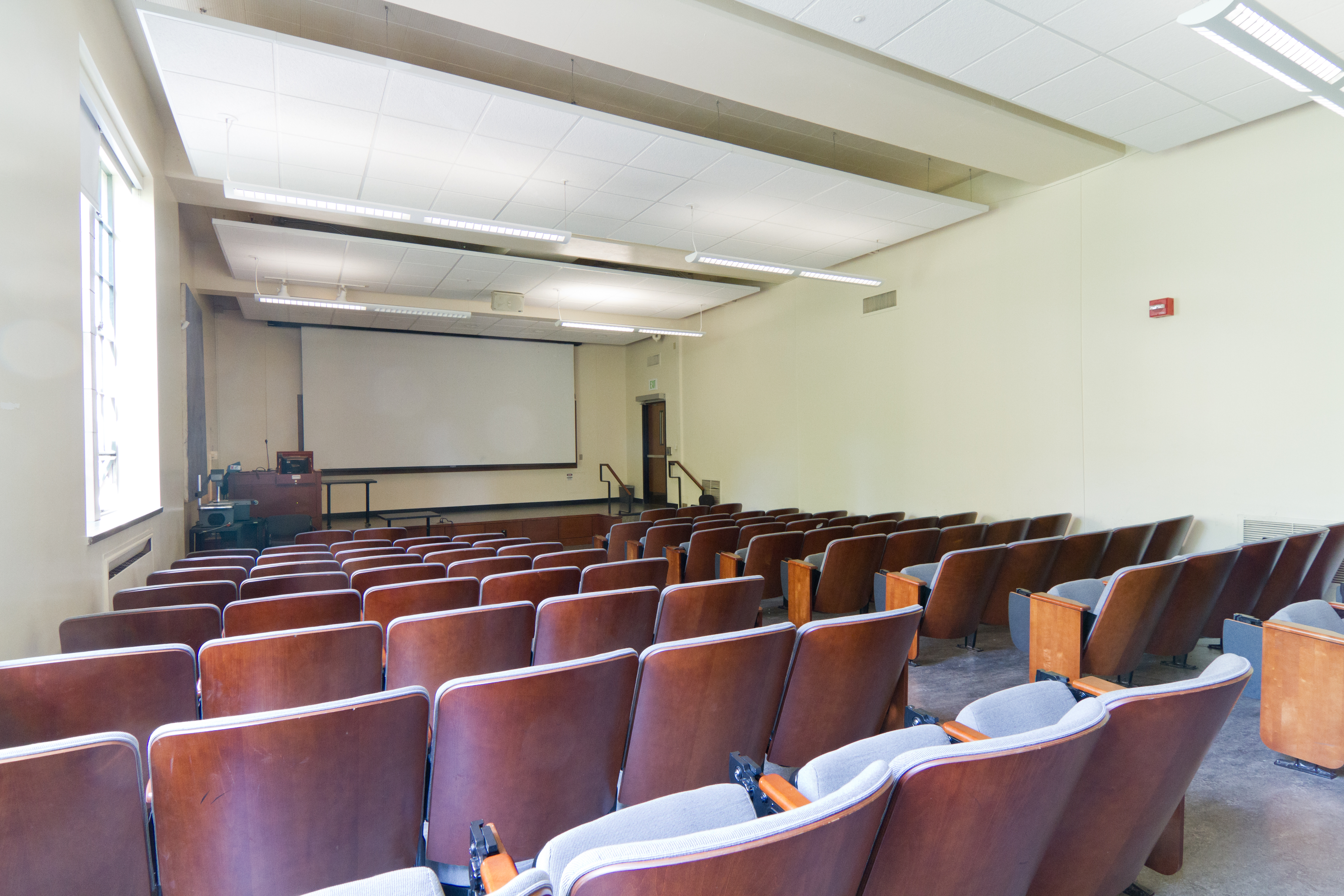 View of rows of chairs facing a lecturn, windows on the left and plain white wall on the right. An aisle slopes downhill without stairs to the lecturn.