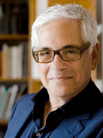 Alexander Nehamas looks into the camera while standing in front of a bookcase and wearing glasses and a collared shirt with a jacket.