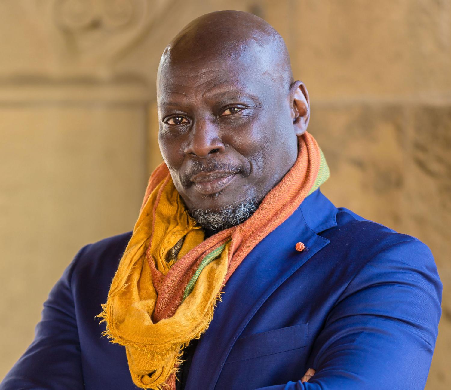 Ato Quayson in a blue suit, orange scarf, smiling at the camera with arms folded.