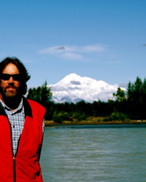 Richard Gray wears a red jacket and stands with a mountain in the background on a clear day.