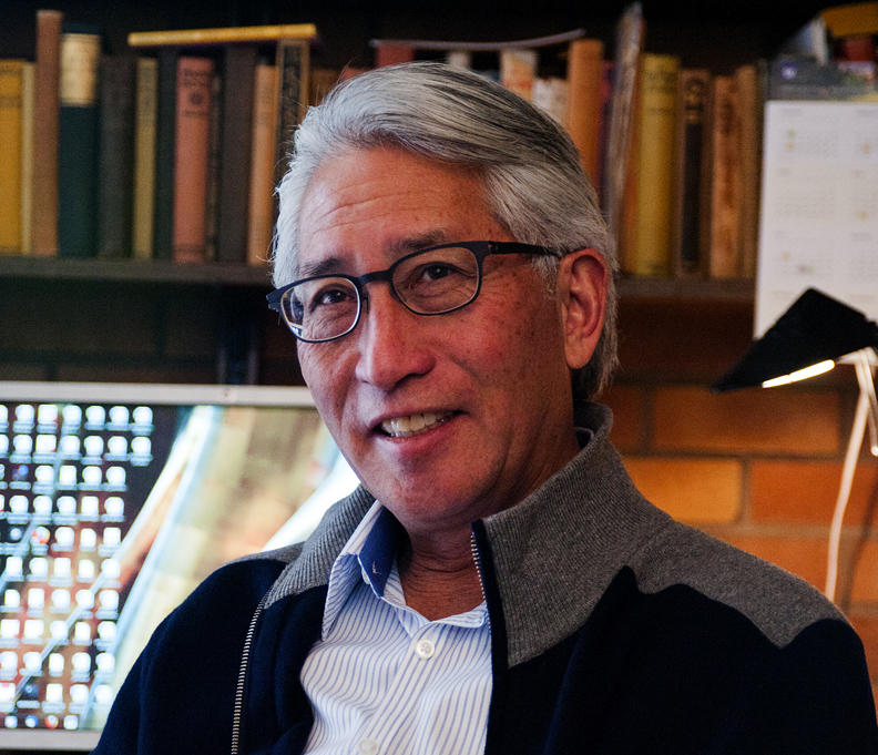 Profile photograph of Shawn Wong. He is Asian and male-presenting with greying short hair, black rimmed glasses, and a smile. He's wearing a shirt under a jacket with bookshelves in the background.