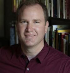 Christopher B. Teuton sits in front of a bookcase wearing a maroon shirt.