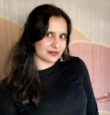 In this photo nanya is wearing a black shirt and standing in front of a beaded curtain and looking at the camera. 
