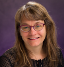 Portrait of Lynn Thomas wearing glasses and sitting in front of a purple wall.