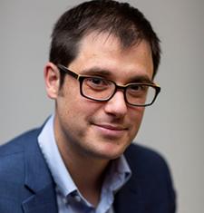 P. Joshua Griffin sits in front of a gray wall wearing glasses, a blue shirt, and a dark jacket.