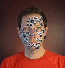 Ted Hiebert sits in front of a dark wall wearing an orange shirt and googly eyes all over his face.