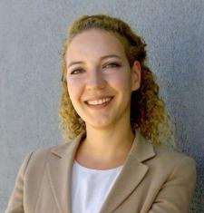 Ayda smiling at the camera in a beige blazer and white shirt, curly hair pulled halfway back and a gray background