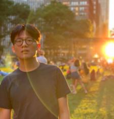 medium close-up of Yandong. He is on the left of the frame in a black t-shirt looking at the camera. To the right is a light flare form the setting sun, while the background shows buildings and a park.