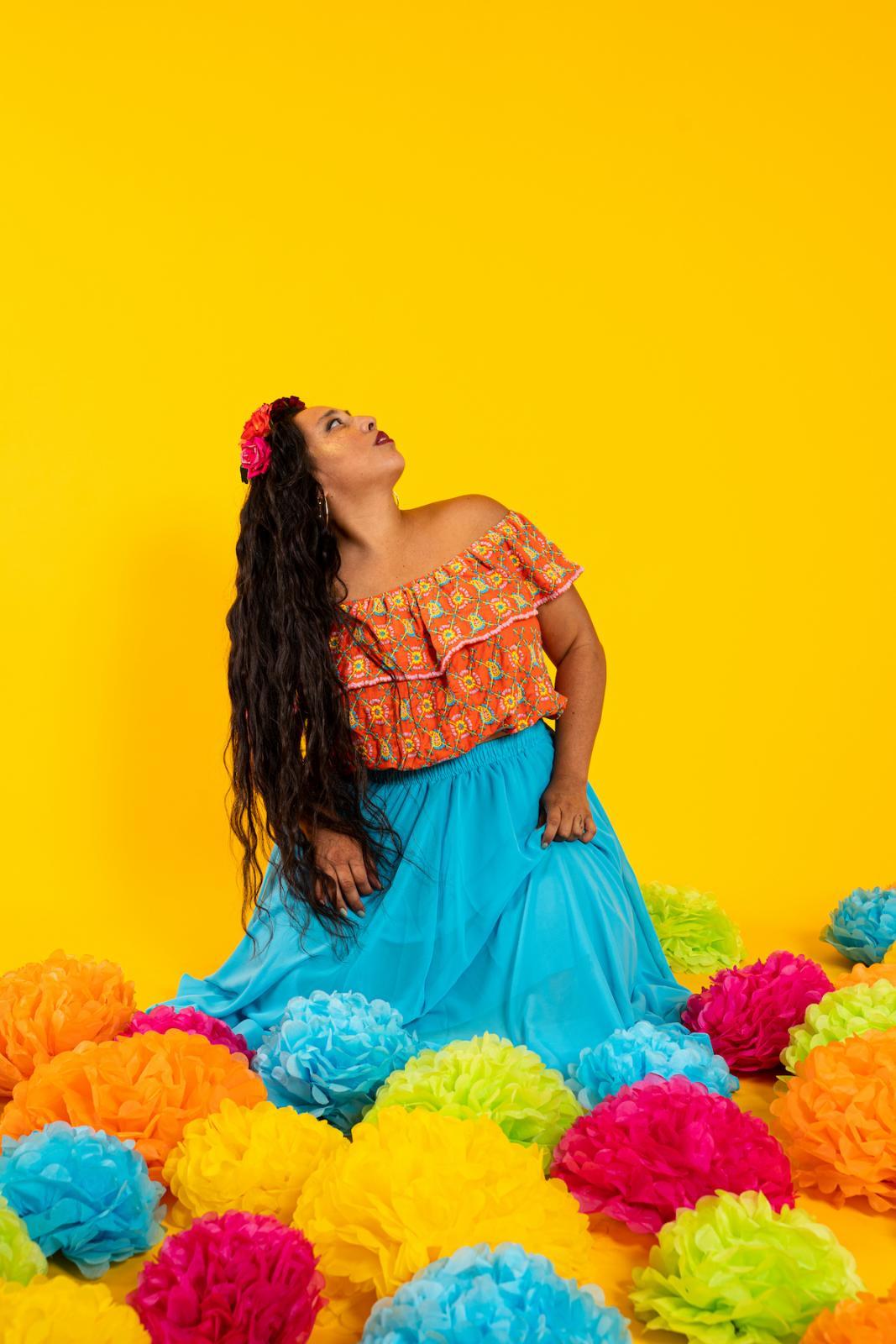 Artist AAlicia Mullikin. She is against a yellow backdrop and surrounded by yellow, green, teal, and bright pink poms on the floor. She has long wavy hair spilling down the side of her shoulder and body and holding a teal dress, looking up to the right. Her top is a print containing the colors of the floor poms and is off the shoulders with ruffles across the top.