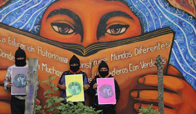 Three people wearing black hats and black masks hold signs while standing in front of a mural depicting a woman with long hair reading a book.