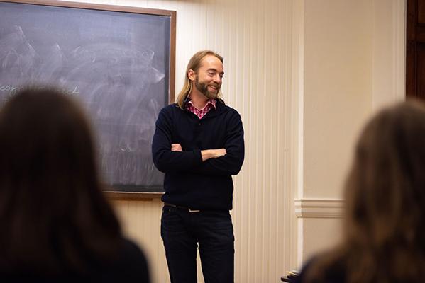 Joel Alden Schlosser stands with arms crossed in front of a chalkboard.