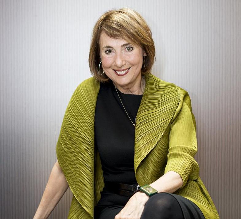 Cathy Davidson smiling at the camera and sitting on a sofa with a green overcoat and black business casual outfit.