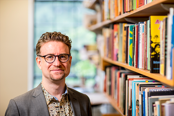 Christian Novetzke in glasses, a grey suit, and a print colored shirt. He is standing next to a line of books on shelves.