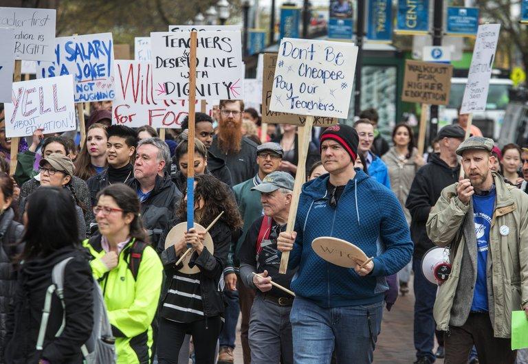 A group of people protest by walking together on a street, holding signs that protest salaries at Seattle-area community colleges.