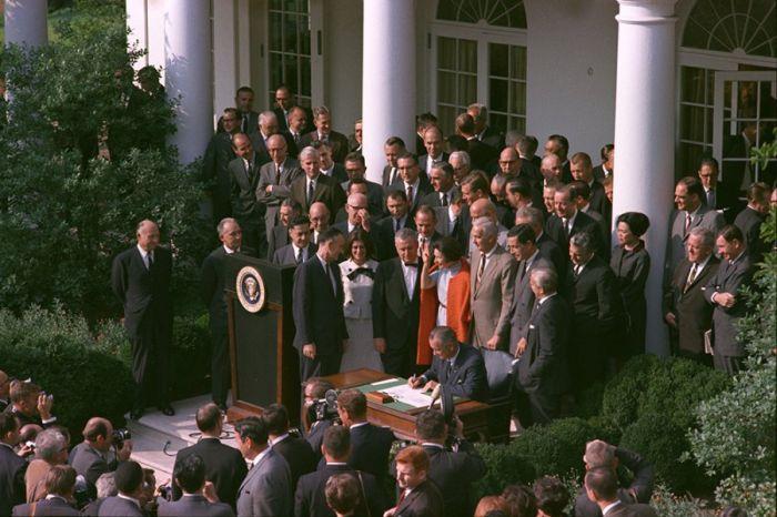 President Lyndon Johnson sits at a desk and signs a document while people in suits stand around him.