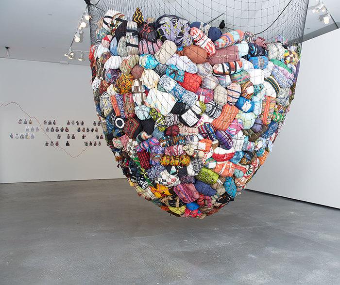Installation art titled Leavings/Belongings by Harriet Bart. A large fishing net hangs from a ceiling, holding bundles of colorful fabric.