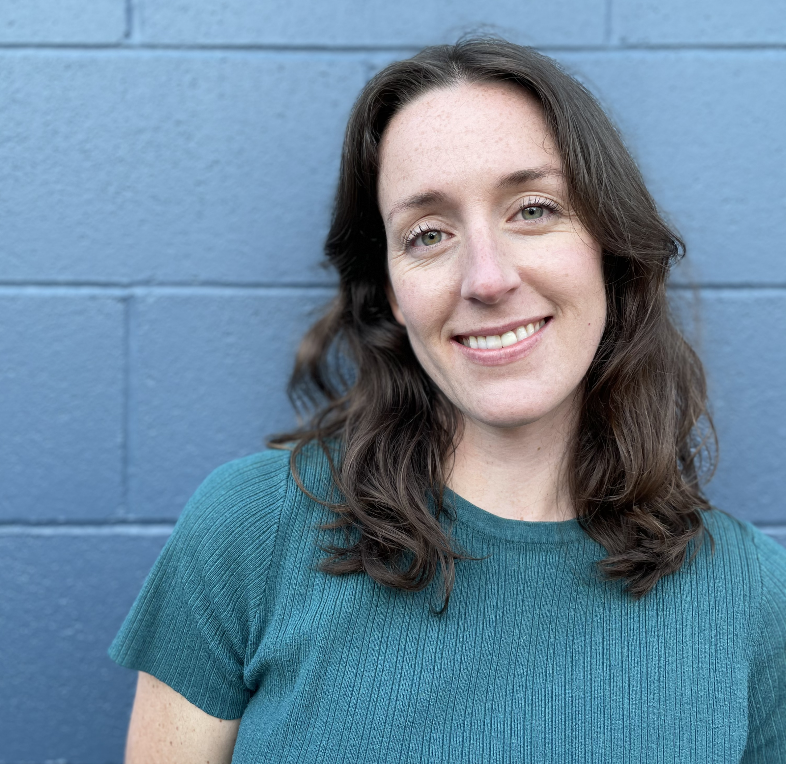 Portrait of Madison Snider: curled, shoulder-length brown hair, blue eyes, smiling at the camera. Blue brick background and wearing a blue t-shirt.