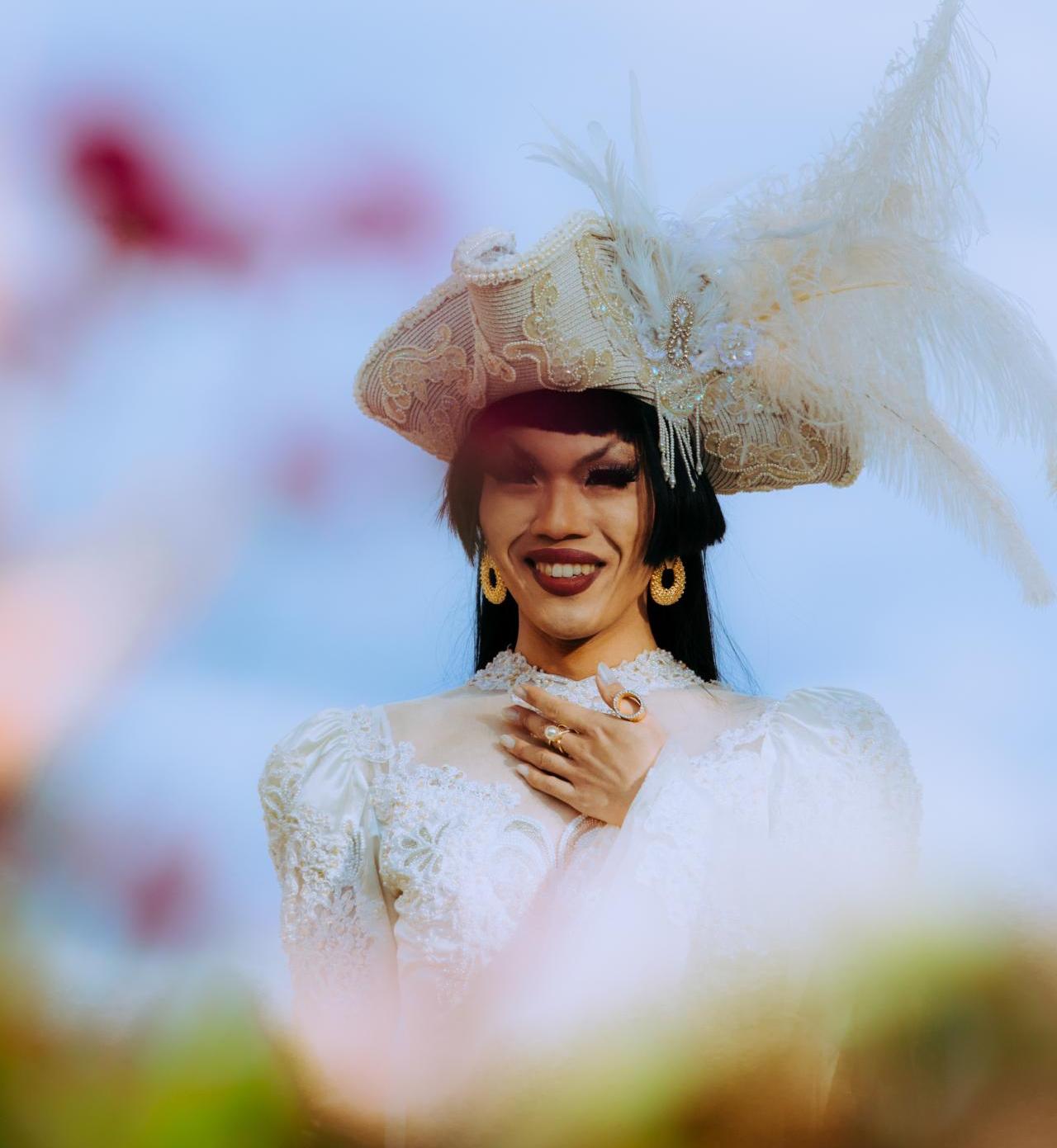 Asian American women in a white hat and dress smiling against a blue sky. Out of focus flowers in the foreground.