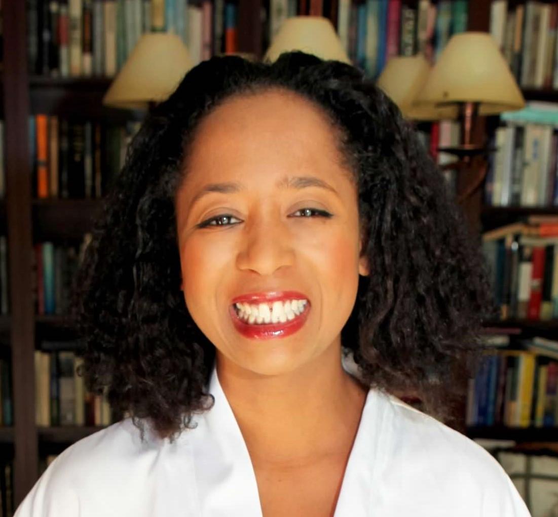 profile photograph of Jasmine Mahmoud: shoulder length curly hair, red lipstick, and a large smile with books in the background