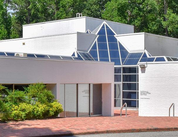 entrance to the national humanities center, which is a white, modern building with angled ceiling windows and a brick courtyard entrance surrounded by green trees and bushes