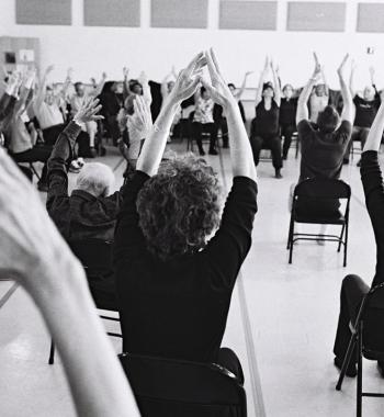 A black and white photo of a group of people from the back sit in chairs with their hands in the air.  