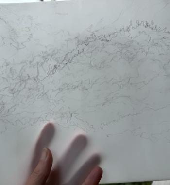 The hand of the artist holding a blind contour drawing of clouds, in pencil on vellum, with a time stamp of 9:07 recorded in the bottom right corner.