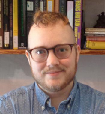 Adrian Kane wears glasses and a blue shirt with a bookcase in the background.