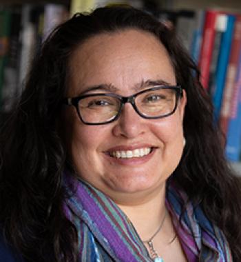 Portrait of Ariana Ochoa Camacho wearing glasses and standing in front of a bookcase.