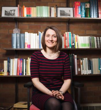 Profile of Beatrice in front of a book case. She has short brown hair and is wearing a sweater while smiling at the camera.