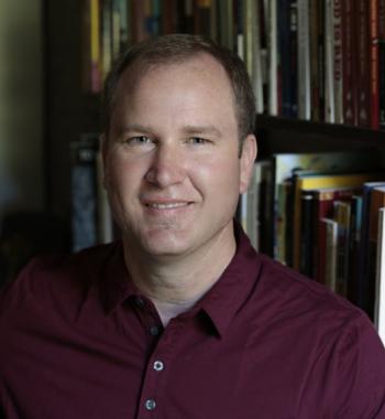 Christopher Teuton sits in front of a bookcase wearing a maroon shirt.