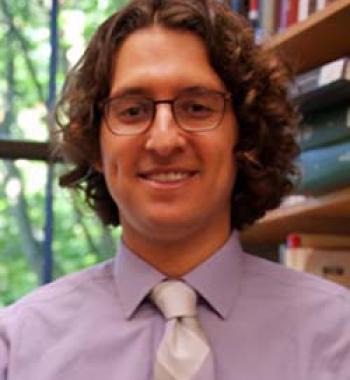 Portrait of Devin Naar wearing a purple shirt and tie and wearing glasses.