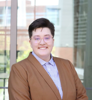 A white butch person with glasses smiling in a light brown blazer and blue and white checkered button-up shirt.