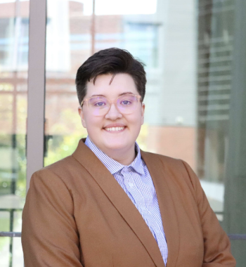 A white butch person with glasses smiling in a light brown blazer and blue and white checkered button-up shirt. 