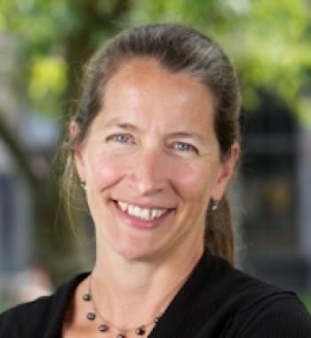 Sara Goering professional photo smiling at the camera, blurred trees in the background, wearing a blue blazer, necklace, and hair pulled back