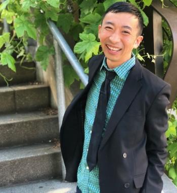 Douglas S. Ishii stands in front of stairs while wearing a blazer, teal collared shirt, and a black tie.