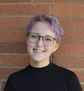 Head and shoulders of a smiling white person with short purple hair and glasses in a black turtleneck against a brick wall background