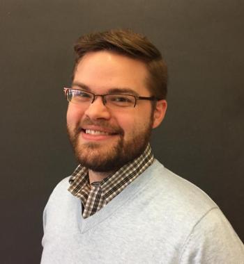 Justin Randolph stands in front of a dark wall while wearing glasses and a white sweater.