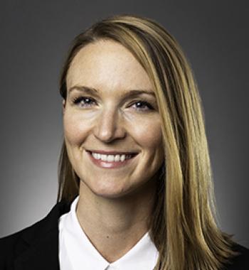 Kristin Dew stands in front of a dark wall wearing a white shirt and dark jacket.