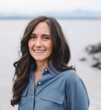 Lauren Drakopulos stands in front of a body of water while wearing a blue button up shirt.