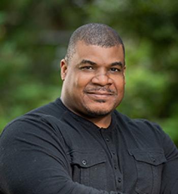 A portrait of Marcus Johnson standing in front of tress wearing a black shirt.