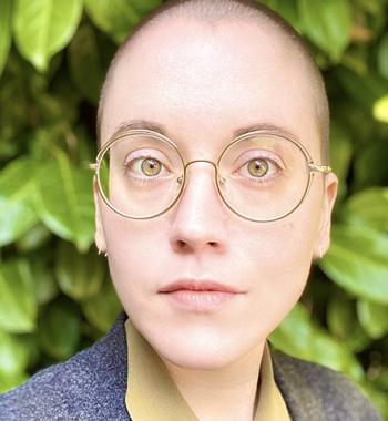 Maxine Savage stands in front of a large bush while wearing glasses and a grey jacket.