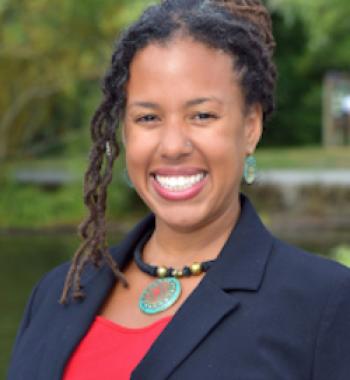 profile of Maya Smith smiling at the camera, hair pulled back with some curls along the left side, wearing a red top under a blue blazer with a necklace